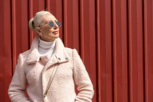 седовласая женщина средних лет в очках на фоне красной металлической стены. Внешний образ шикарной дамы средних лет в солнцезащитных очках и светлых волосах, наслаждающейся прекрасным летним днем, в розовой куртке - Фото, изображение