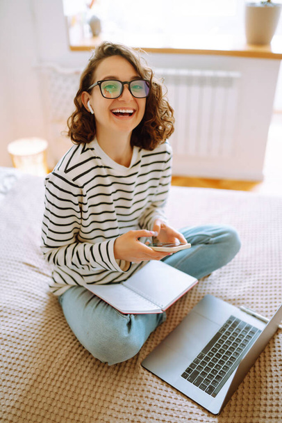 Education and working online. Young female freelancer sitting at comfortable bed in cosy home interior and working remotely on modern laptop computer. Online shopping in web store. - Foto, Imagem
