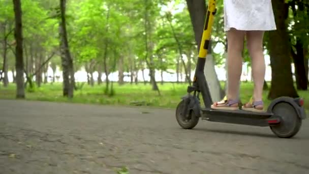 Jonge vrouw in een witte jurk rijdt een delende electro scooter in het park. Onderaanzicht. - Video
