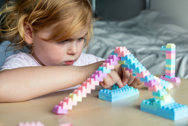 Kis gyönyörű gyermek játszik játék műanyag építőkockák, ül az asztalnál. Kislány elfoglalt szórakoztató kreatív szabadidős tevékenység. Finom motoros készségek fejlesztése gyermekek számára. - Fotó, kép