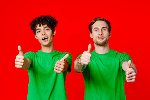 Amigos alegres en camisetas verdes haciendo gestos con las manos emociones fondo rojo - Foto, imagen