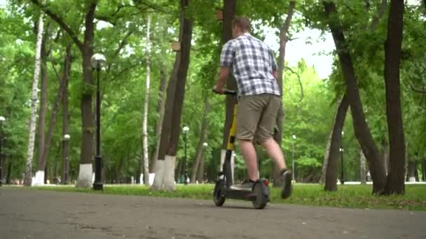 Een jonge Europeaan in overhemd en korte broek rijdt op een elektrische scooter in het park. Onderaanzicht. - Video