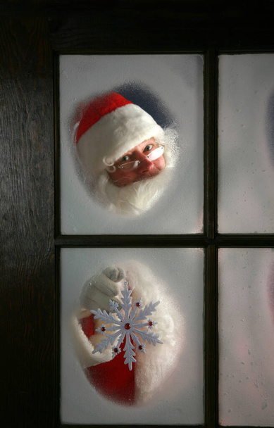 Le Père Noël regarde à travers la neige et le brouillard sur la fenêtre de son atelier au pôle nord pour voir à l'extérieur vérifier la météo tandis qu'il tient un grand flocon de neige le 24 décembre, veille de Noël - Photo, image