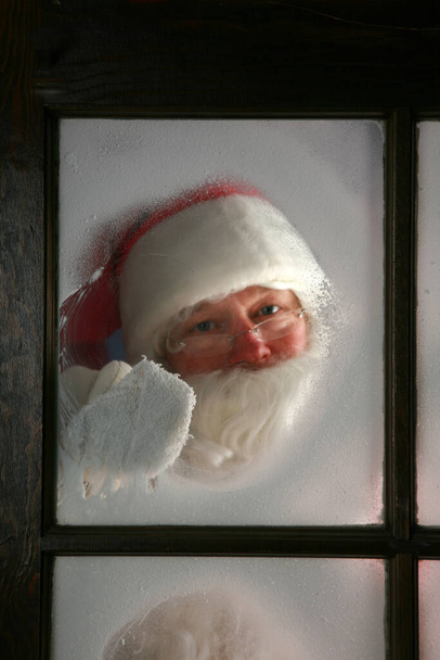 Ο Άγιος Βασίλης σκουπίζει το χιόνι και την ομίχλη μακριά από το παράθυρο του εργαστηρίου του στο βόρειο πόλο για να δει έξω και να βεβαιωθεί ότι οι τάρανδοι του ετοιμάζονται να πάνε για δουλειά στις 24 Δεκεμβρίου, παραμονή Χριστουγέννων. - Φωτογραφία, εικόνα