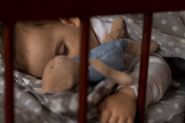 Мила весела маленька пухка дівчинка, яка солодко спить у сірому дитячому ліжечку під час обіду відпочиває в біло-рожевій піжамі з плюшевим ведмедем вдома. Дитинство, дозвілля, комфорт, медицина, концепція здоров'я
 - Фото, зображення