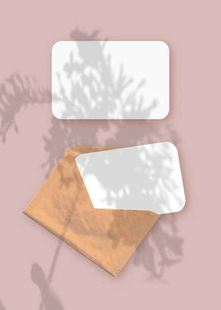 Макет с наложением тени растения на конверт с двумя листами текстурированной белой бумаги на розовом фоне стола. Вертикальная ориентация - Фото, изображение