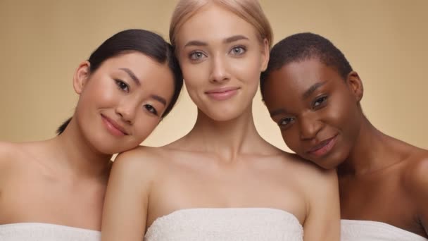 Tres jóvenes mujeres multiétnicas posando envueltas en toallas sobre fondo beige del estudio, abrazando y sonriendo a la cámara - Imágenes, Vídeo