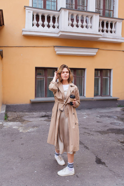 Young millennial woman with wild hair dressed in an autumn coat walking with a cup of coffee to go near the old building. - Фото, зображення