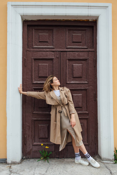 Jeune femme millénaire aux cheveux sauvages vêtue d'un manteau d'automne posant près de la porte d'un vieux bâtiment. - Photo, image