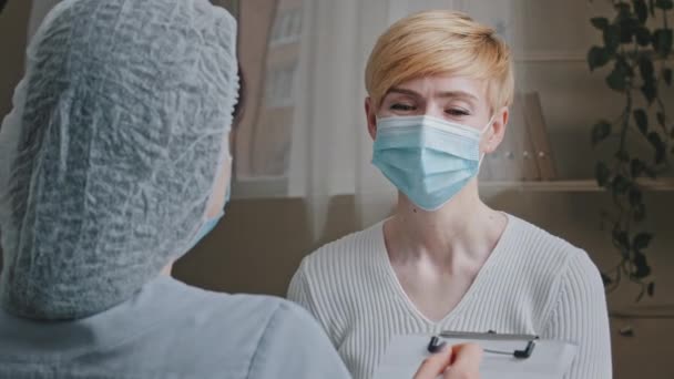 View from behind unrecognizable woman doctor specialist practitioner medical worker consults mature sick female patient in protective mask asks about symptoms disease writes down diagnosis in hospital - Filmagem, Vídeo