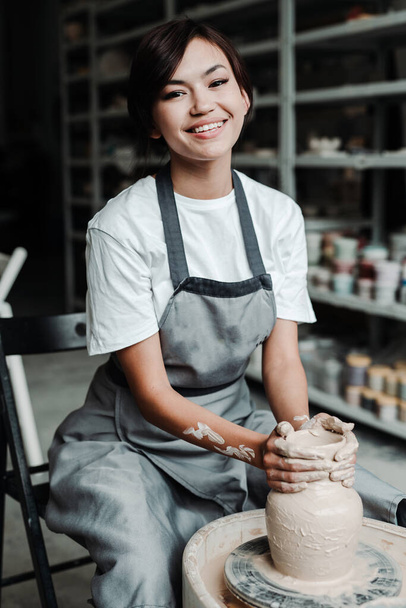 Una donna asiatica carina si siede a una ruota vasai in uno studio hobby e fa un vaso di argilla. Bel sorriso, denti bianchi - Foto, immagini