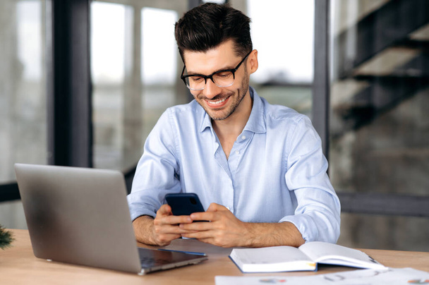 Joyful caucasian young man, manager or freelancer sitting at his desk in the office, texting with a friend or client using a smartphone, online chat, browsing the Internet, answering email, smiling - Fotó, kép