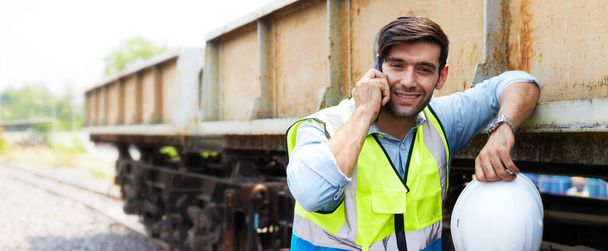 Un ingeniero ferroviario o un técnico de mantenimiento en ropa protectora se está relajando durante el descanso sonriendo en el teléfono cerca de un viejo tren de carga.. - Foto, imagen