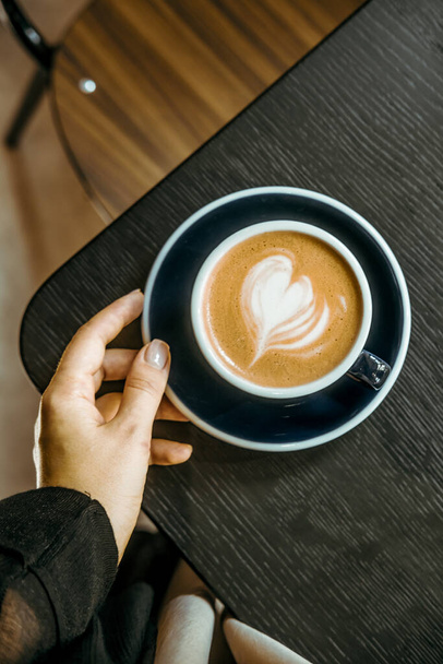 Ręka kobiety trzymającej kawę cappuccino na czarnym drewnianym stole w kawiarni. Koncepcja fotografii napojów spożywczych. Uroczy, spokojny, smaczny, nastrojowy strzał. Blisko, widok z góry - Zdjęcie, obraz