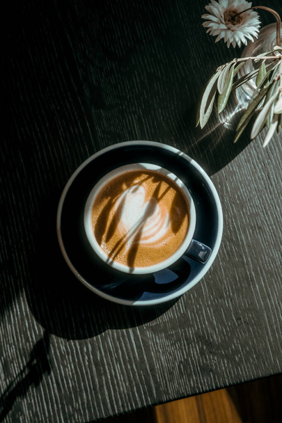 Cappuccino-Kaffee auf schwarzem Holztisch im Café. Licht und Schatten, Food Drink Fotografiekonzept. Schöner, ruhiger, schmackhafter, stimmungsvoller Stimmungsschuss. Nahaufnahme, Draufsicht - Foto, Bild