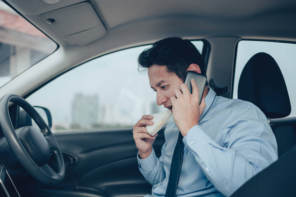 Businessman eating sandwich while working in laptop and talking on the phone  in the drivers seat in his car. Busy businessman and food in car - Foto, Imagen