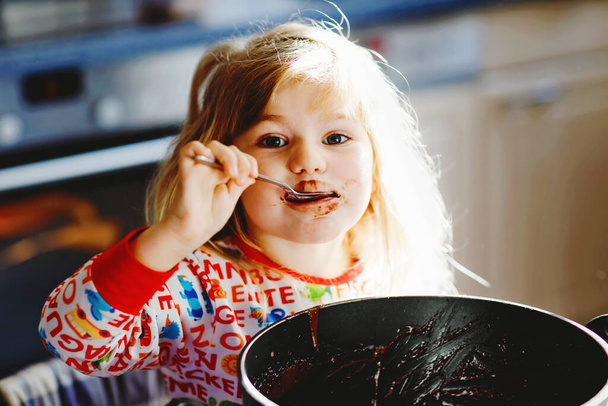 Cute toddler girl eating chocolate dough rests with spoon and fingers from pot. Happy child licking sweet dough for muffins or cake, helping in home kitchen, indoors. - Фото, изображение