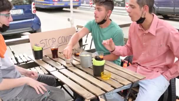 Drie vrienden in een buitenruimte, op de tafel een bord met de woorden stop covid. Ze hebben maskers laten zakken om te kunnen drinken. Begrip vriendschap, nieuw normaal, strijd tegen de coronaviruspandemie. - Video