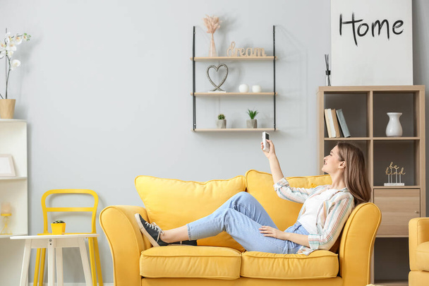 молодая женщина с пультом управления кондиционером сидит на диване в гостиной - Фото, изображение