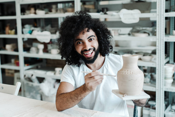  A curly-haired guy makes patterns on a clay vase in a pottery workshop. Arab oriental appearance - Foto, Imagen