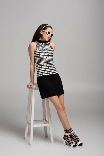 full length of young woman in retro outfit and sunglasses leaning on high stool while posing on grey  - Foto, Bild