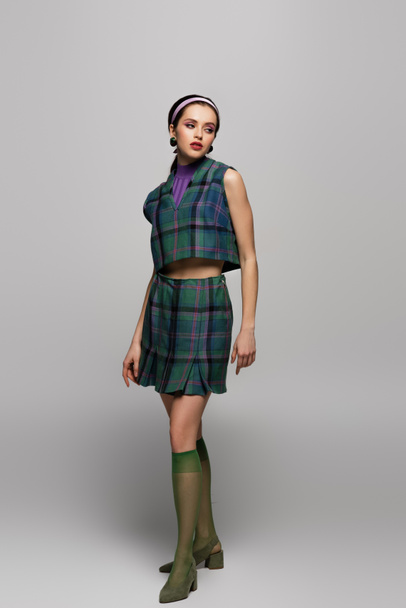 full length of young model in headband and vintage checkered outfit posing on grey  - Foto, Imagen