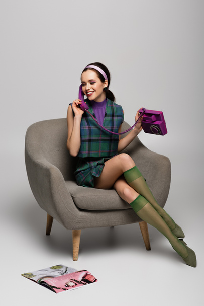 happy young woman talking on retro phone while sitting in armchair near magazine on floor on grey - Photo, Image