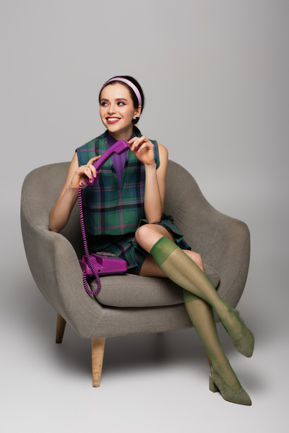 pleased young woman holding purple retro phone while sitting in armchair on grey - Foto, Imagem