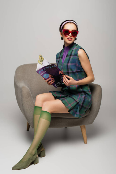 young woman in retro sunglasses holding magazine while sitting in armchair on grey - Foto, imagen