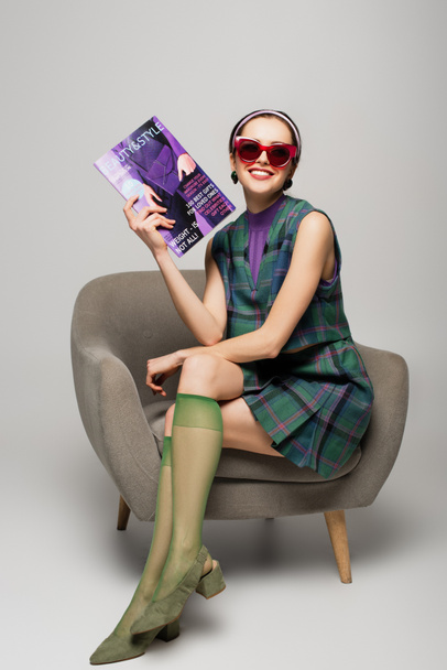 positive young woman in sunglasses holding magazine while sitting in armchair on grey - Foto, imagen