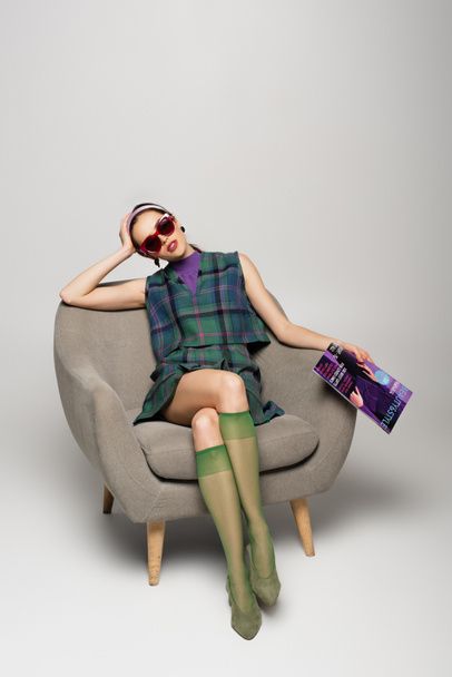 young woman in sunglasses holding magazine while sitting in armchair on grey - Foto, afbeelding