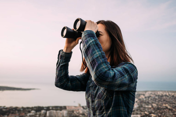 Portrait of a young female tourist looking through binoculars at sunset. - 写真・画像