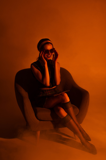 glückliche junge Frau setzt Sonnenbrille auf und sitzt im Sessel mit Magazin auf Orange - Foto, Bild