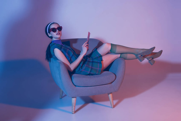 brunette young woman in sunglasses and retro outfit sitting in armchair with magazine on violet - Φωτογραφία, εικόνα