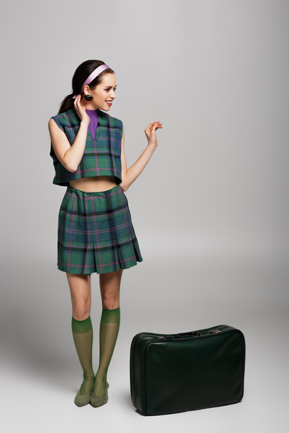 longitud completa de la mujer feliz en traje retro de pie cerca de la maleta verde en gris - Foto, Imagen