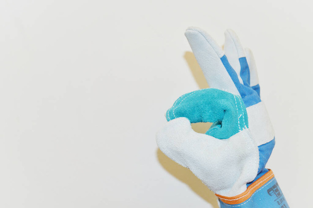 Hand in blue work cloth glove shows ok sign against white wall background - Foto, Imagen