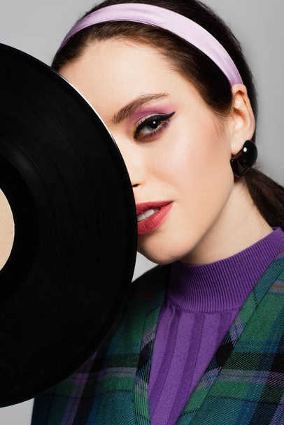 young woman in headband looking at camera near retro vinyl disc isolated on grey - Fotografie, Obrázek