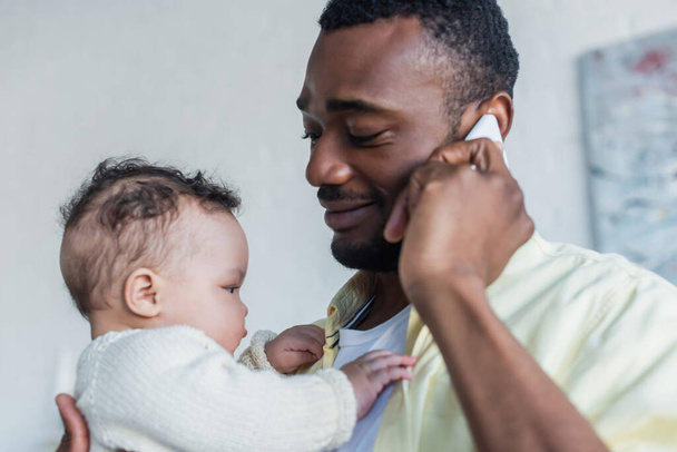 улыбающийся африканский американец разговаривает по смартфону, держа в руках новорожденную дочь - Фото, изображение