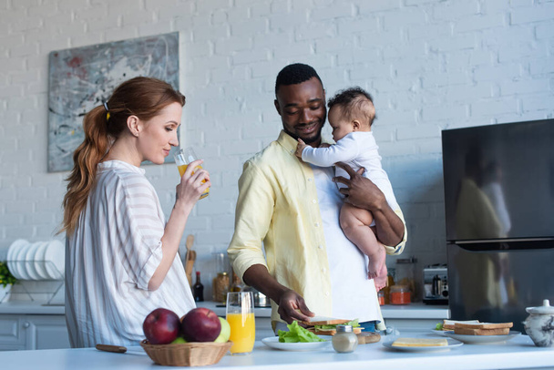 africano americano hombre con bebé chica preparación sándwiches cerca esposa beber naranja jugo - Foto, imagen