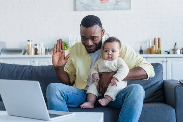 幸せなアフリカ系アメリカ人男性と小さな娘手を振ってビデオ通話中にノートパソコンで - 写真・画像