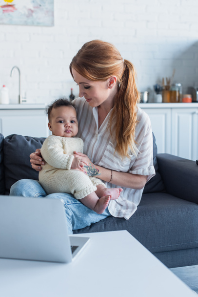 woman sitting on couch with infant african american daughter near laptop - Fotó, kép