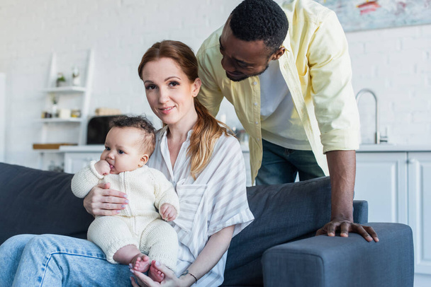 african american man looking at happy wife sitting on sofa with infant daughter - Foto, Bild