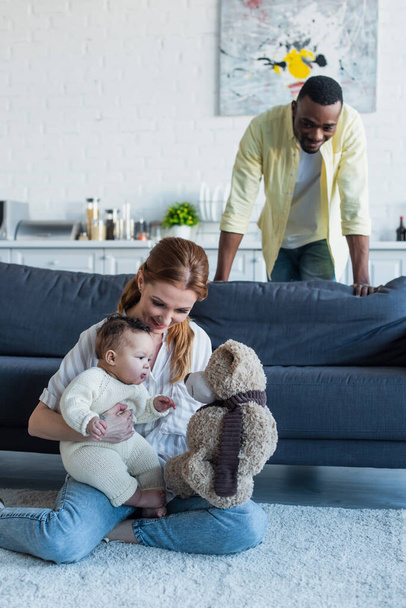 happy african american man looking at wife playing with daughter and teddy bear on floor - Фото, зображення