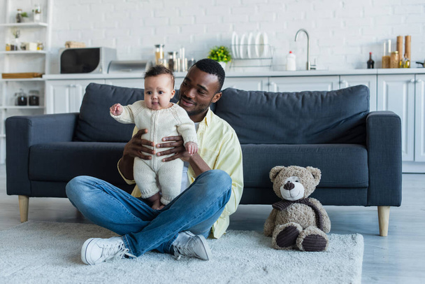 smiling african american man holding infant kid while sitting on floor near sofa and teddy bear - Фото, изображение