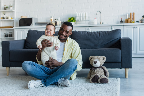 cheerful african american man sitting on floor with excited baby near teddy bear - Photo, Image