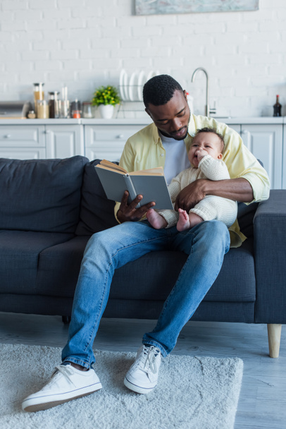 homme afro-américain assis sur le canapé avec bébé et livre de lecture - Photo, image
