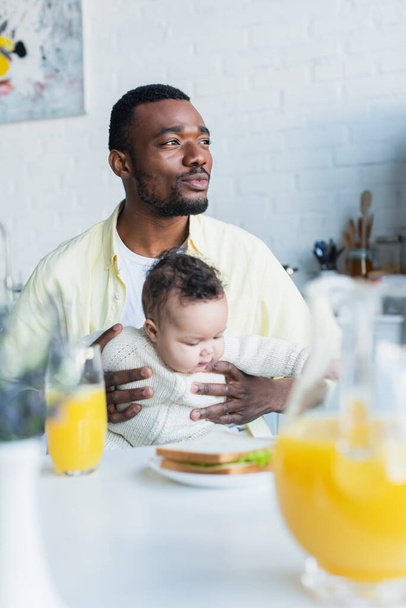 africano americano hombre mirando hacia otro lado mientras sentado con bebé cerca borrosa tarro de jugo de naranja - Foto, Imagen