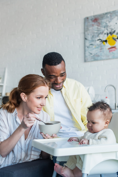 happy woman feeding infant kid near african american husband - Valokuva, kuva