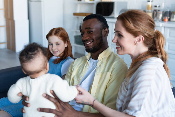 szczęśliwy międzyrasowy rodzina patrząc na niemowlę dziewczynka siedząc w domu - Zdjęcie, obraz