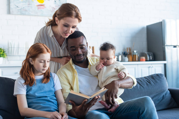 Afrykański Amerykanin czyta książkę wielonarodowej rodzinie na kanapie w domu - Zdjęcie, obraz